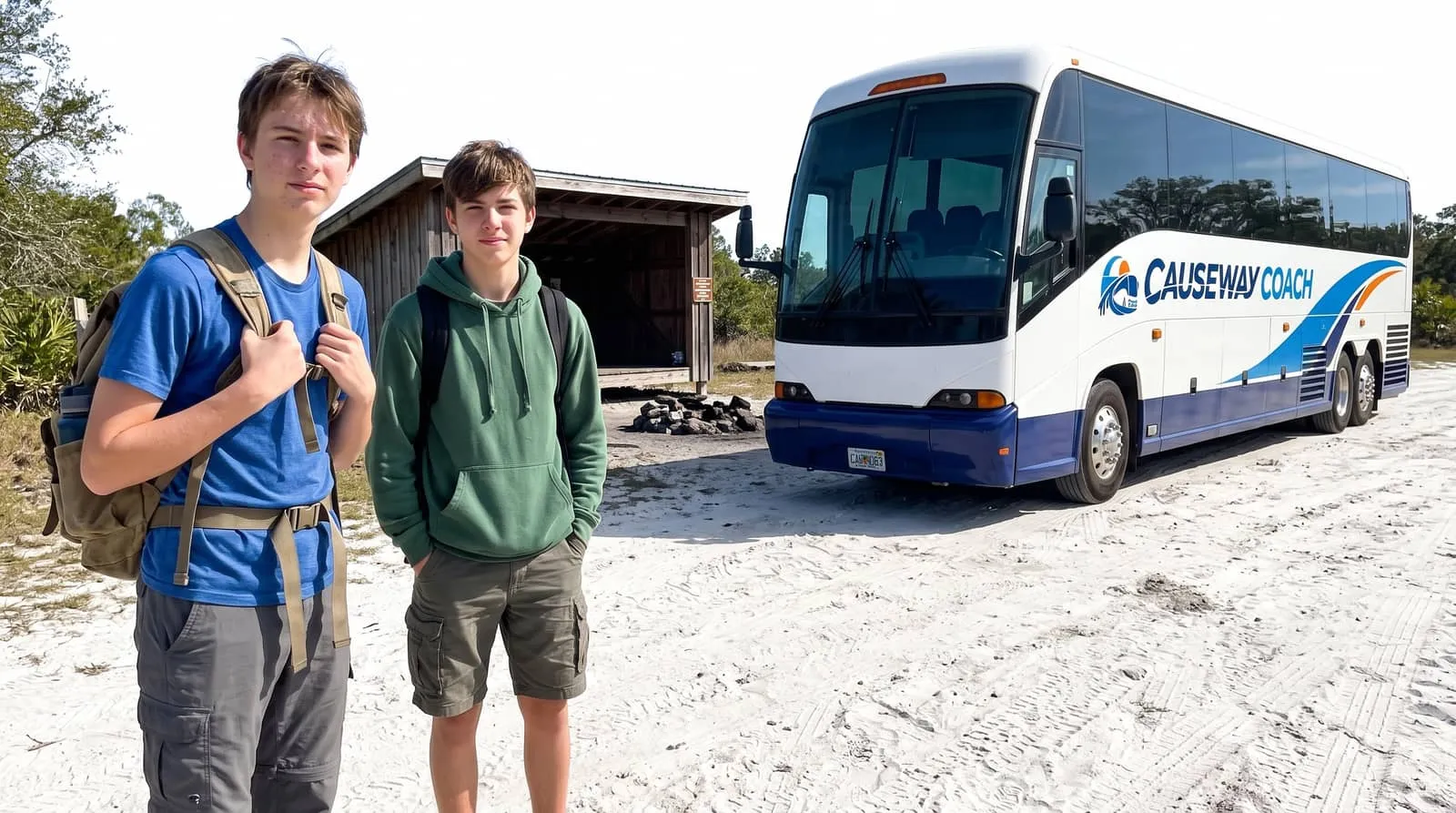 Two teenagers with daypacks beside a Causeway Coach motorcoach on a sandy track at a scout camp in Volusia County
