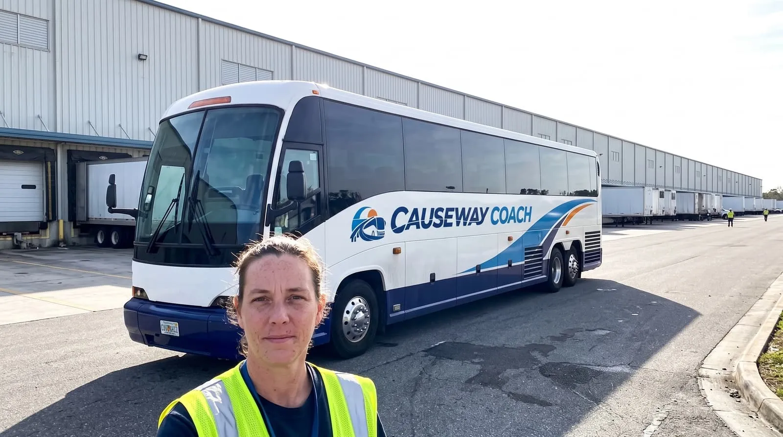A warehouse worker in a hi-vis vest at the door of a Causeway Coach shuttle beside a large distribution centre in north Jacksonville